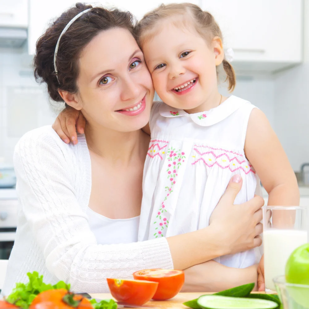 About 2 Smiling mother and daughter hugging in a bright kitchen with fresh vegetables on the table.